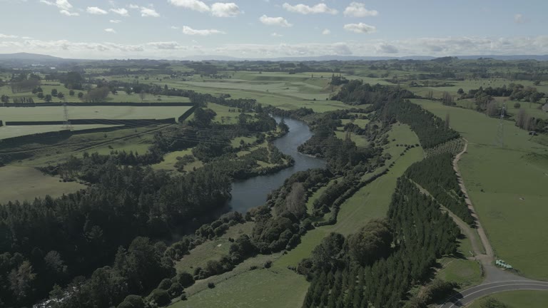 An aerial view follows a winding river between green fields, trees and low hills.